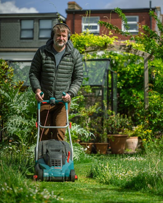 A man with short, light brown hair and a beard is performing garden maintenance in a lush backyard during daytime. He is wearing a black puffer jacket over a gray shirt and brown trousers, and is usin