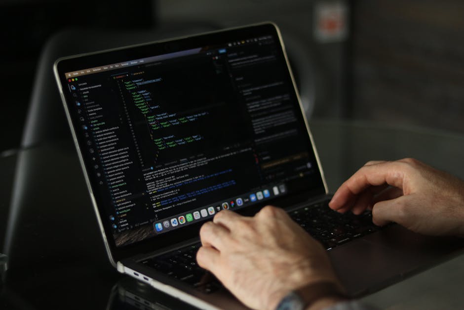A person working on a laptop placed on a dark, polished table, with visible code editors and programming scripts displayed on the screen. The individual's hands are positioned over the keyboard, activ